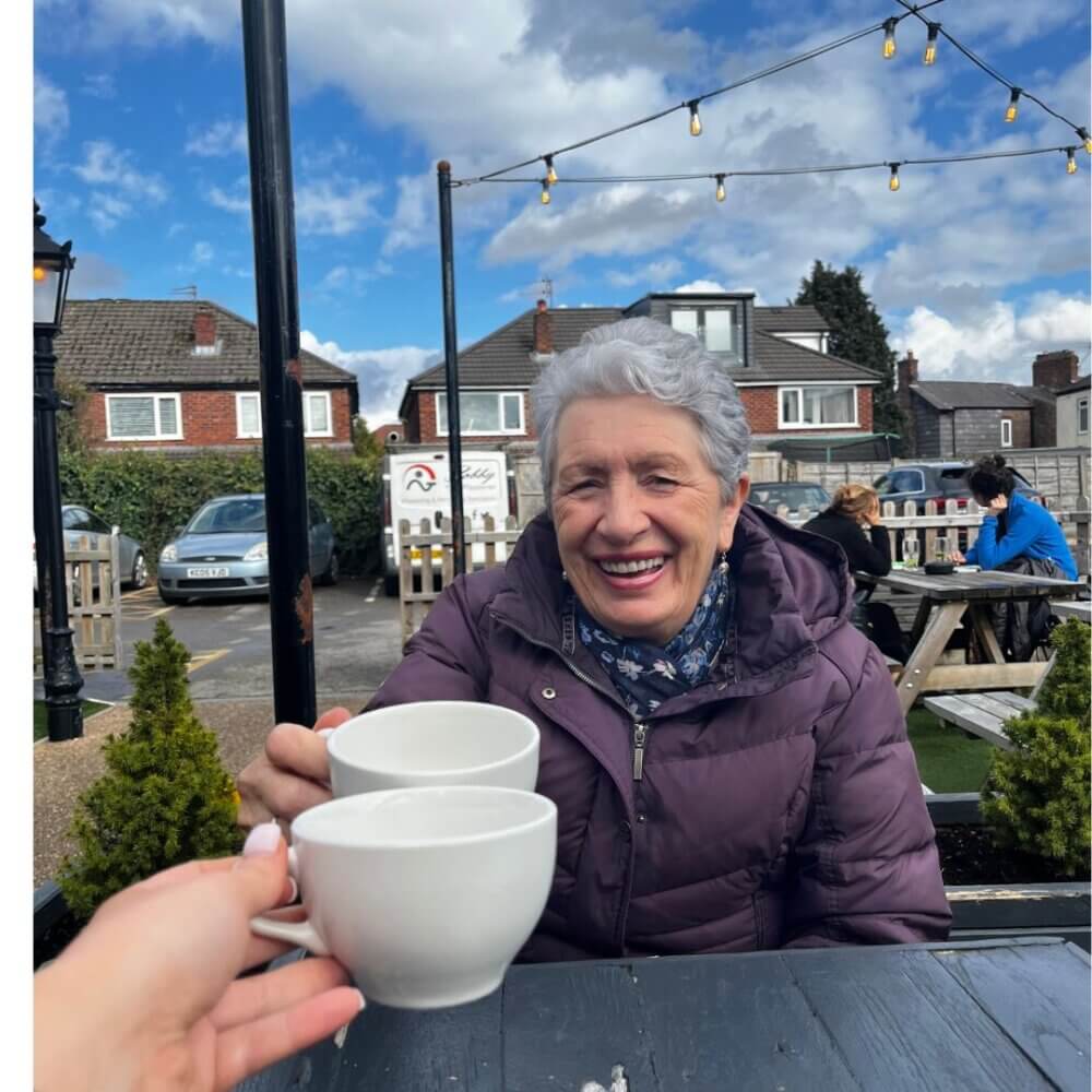 A smiling elderly woman in a purple jacket holds a cup, clinking it with another cup at an outdoor café with string lights. - Home Instead
