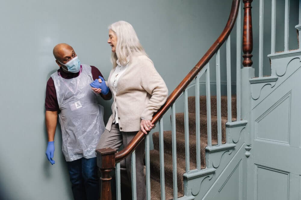A caregiver helps an elderly woman down the stairs while holding her arm and wearing a mask, gloves, and an apron. - Home Instead
