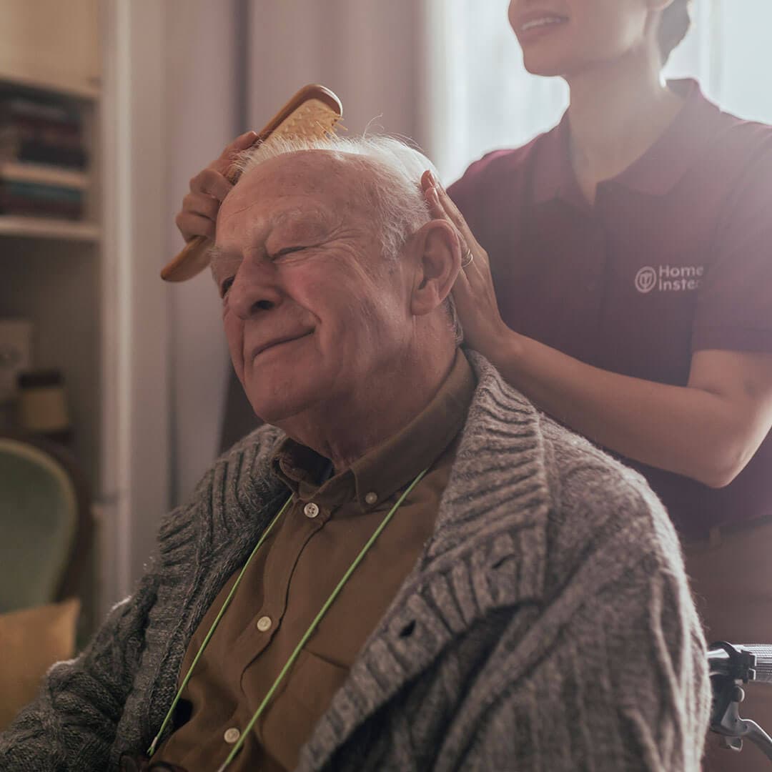 Elderly man enjoying a caregiver from "Home Instead" brushing his hair, both smiling in a cozy home setting. - Home Instead