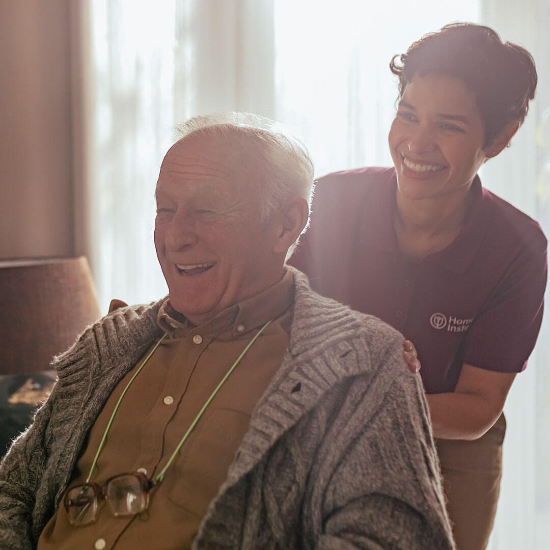 Elderly man sits and laughs with a caregiver in uniform who is gently touching his shoulders, both smiling warmly. - Home Instead
