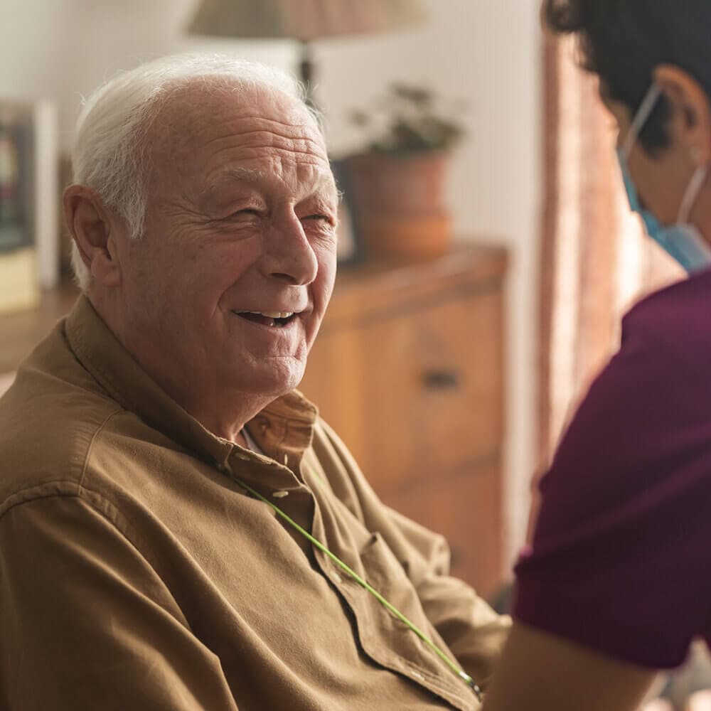 An elderly man smiling while a caregiver in a mask and purple shirt tends to him in a warmly lit room. - Home Instead