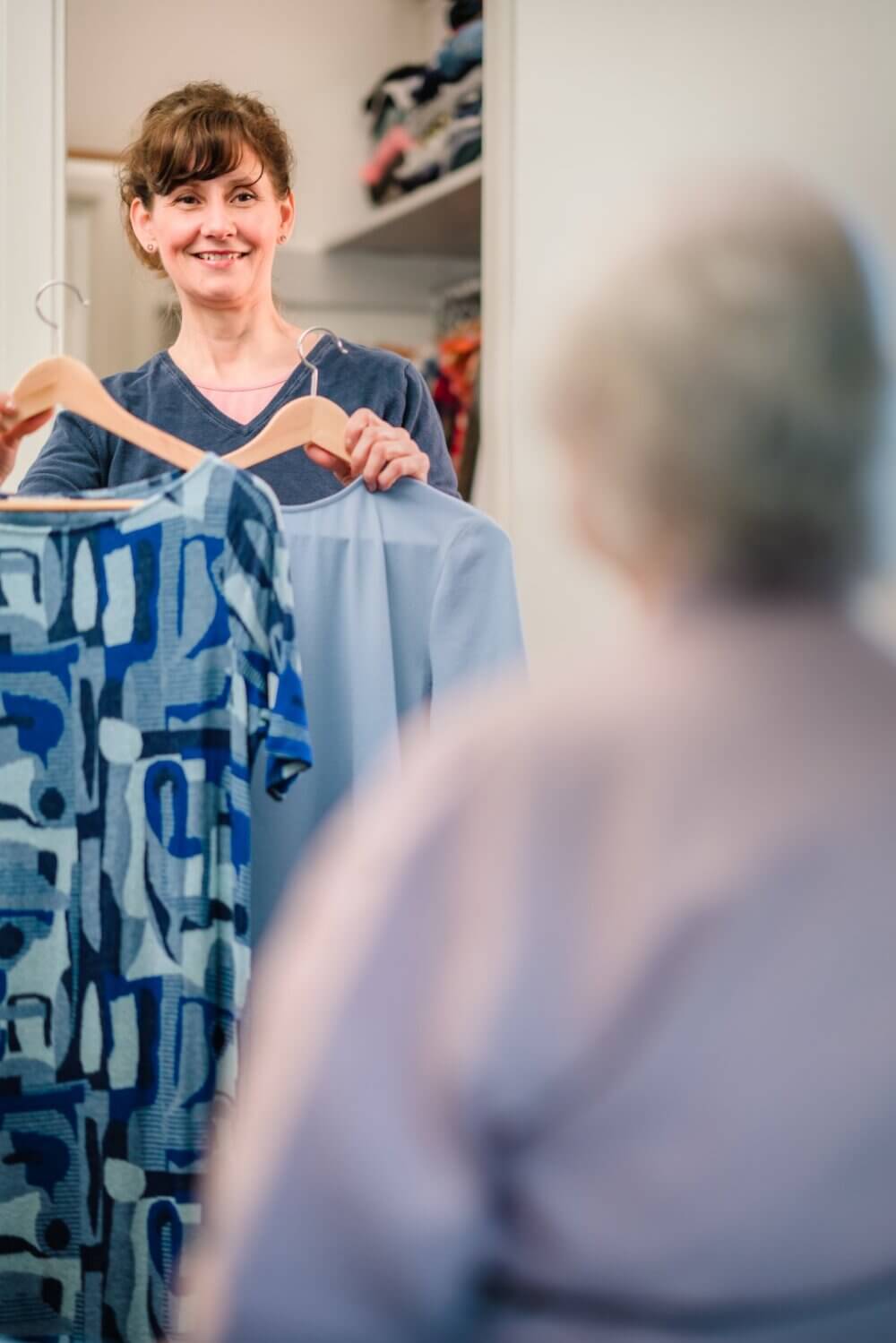 Person holding up two clothing options, smiling at another person sitting in front of them. Closet with clothes in background. - Home Instead