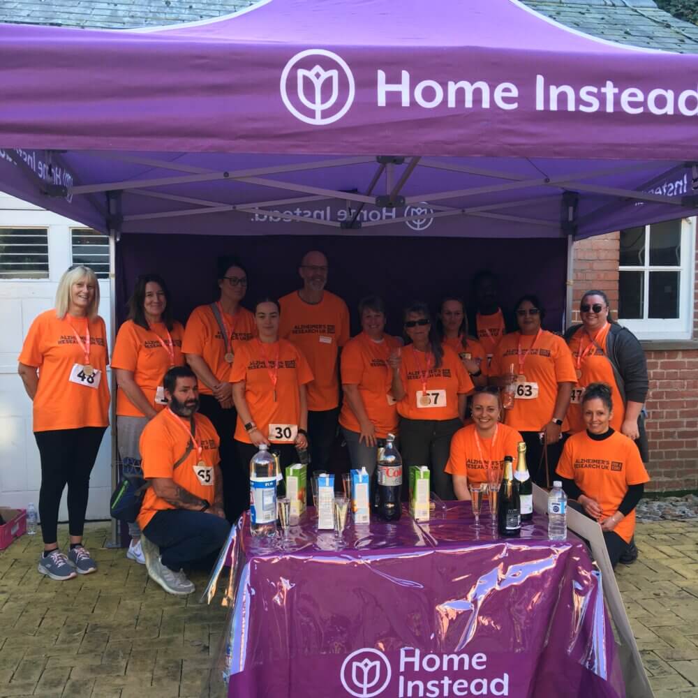 A group of people in orange shirts pose under a Home Instead tent at an outdoor event, with a table of refreshments in front. - Home Instead