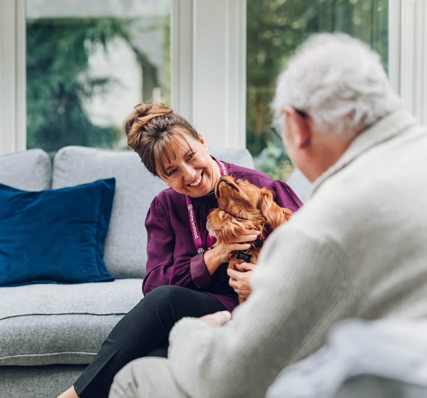 A woman smiling and holding a dog while sitting on a couch, talking to an elderly man in a sunny room. - Home Instead