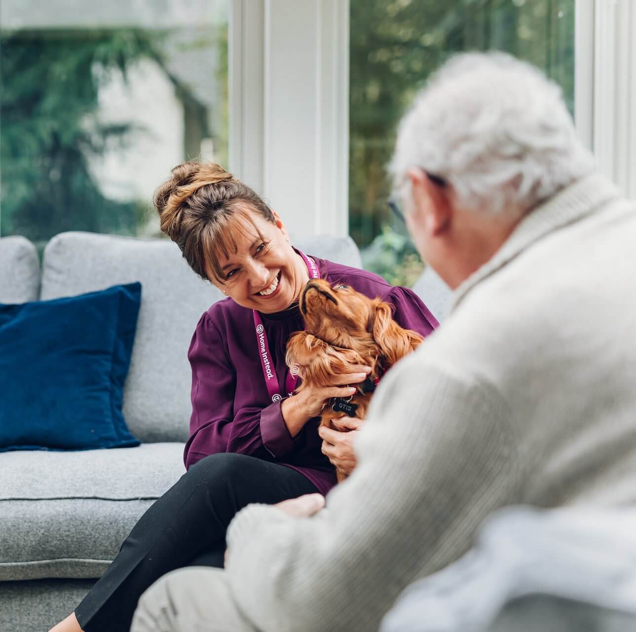 A woman with a dog sits on a sofa, smiling at an elderly person seated across from her. - Home Instead