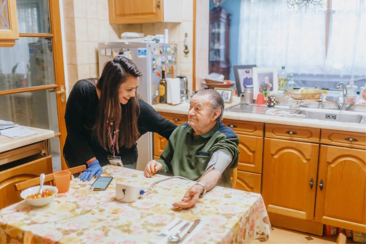 A woman assists an elderly man with a blood pressure monitor at a kitchen table. Both are smiling and talking. - Home Instead