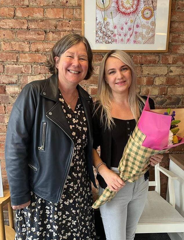 Two women standing together smiling, one holding a bouquet of flowers, with a brick wall and floral artwork behind them. - Home Instead