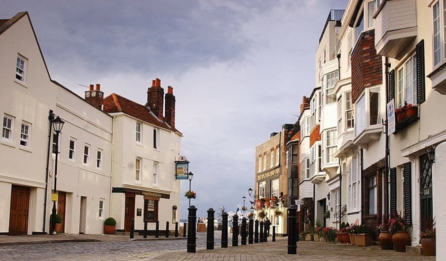 A narrow, cobblestone street lined with quaint buildings under a cloudy sky, leading toward a waterfront. - Home Instead