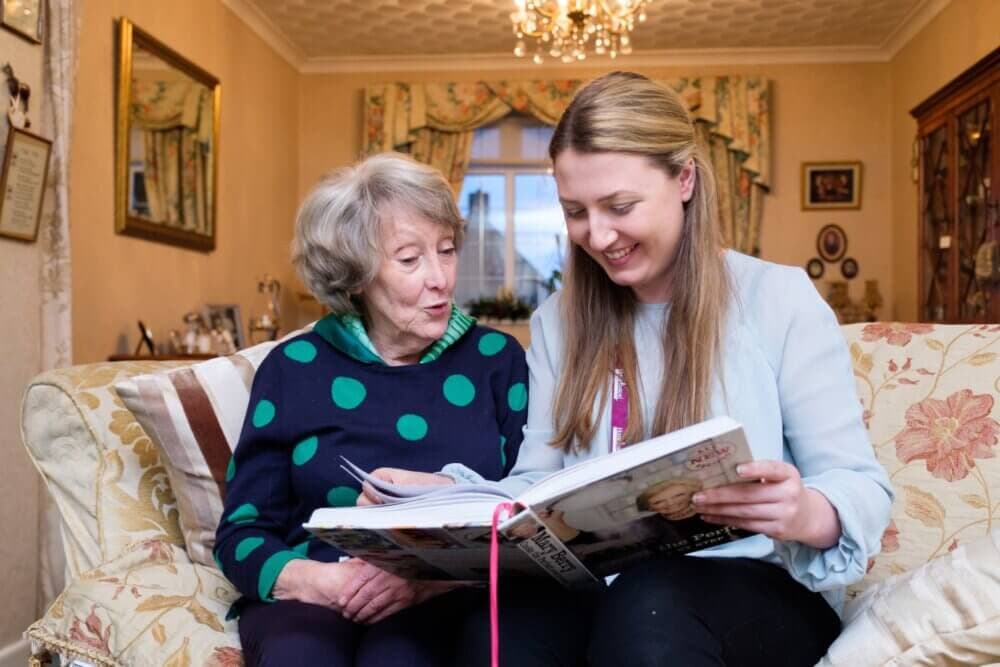 Two women sitting on a couch, smiling and looking at a large open book together in a cozy, well-decorated room. - Home Instead