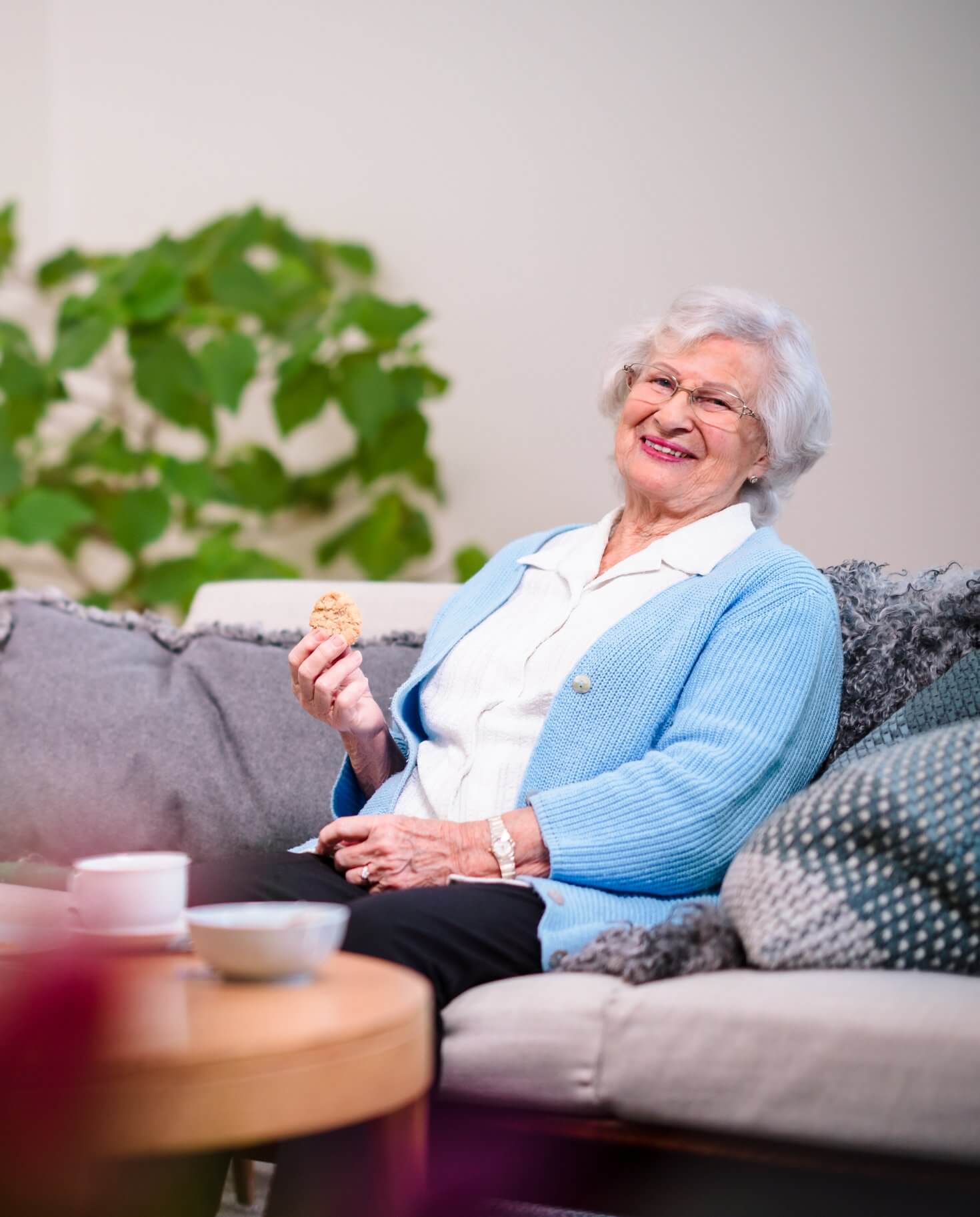 An elderly woman with white hair, wearing a blue cardigan, sits smiling on a couch, holding a cookie. - Home Instead