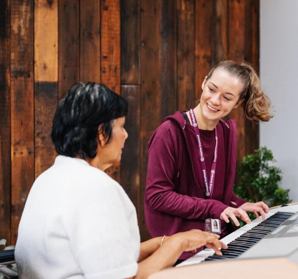 Two women, one elderly and one young, smiling and playing a keyboard together in a wooden-paneled room. - Home Instead
