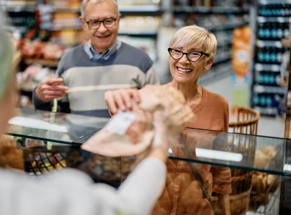 Two smiling elderly people receiving sliced meat at a grocery store deli counter. - Home Instead