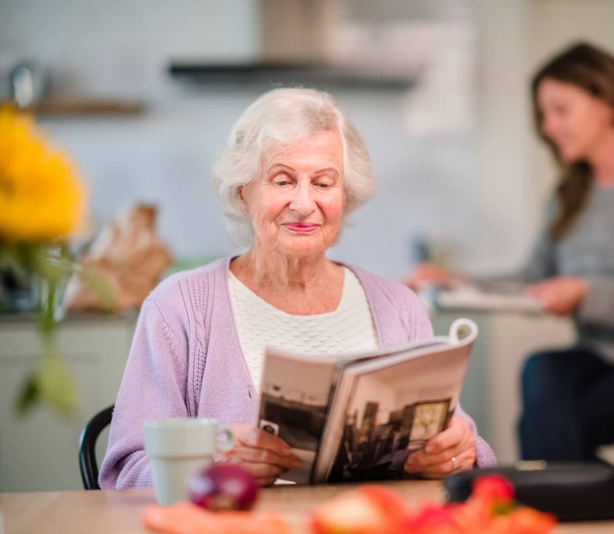 Elderly woman reading a magazine at a table with fruit, while another person sits in the background. - Home Instead