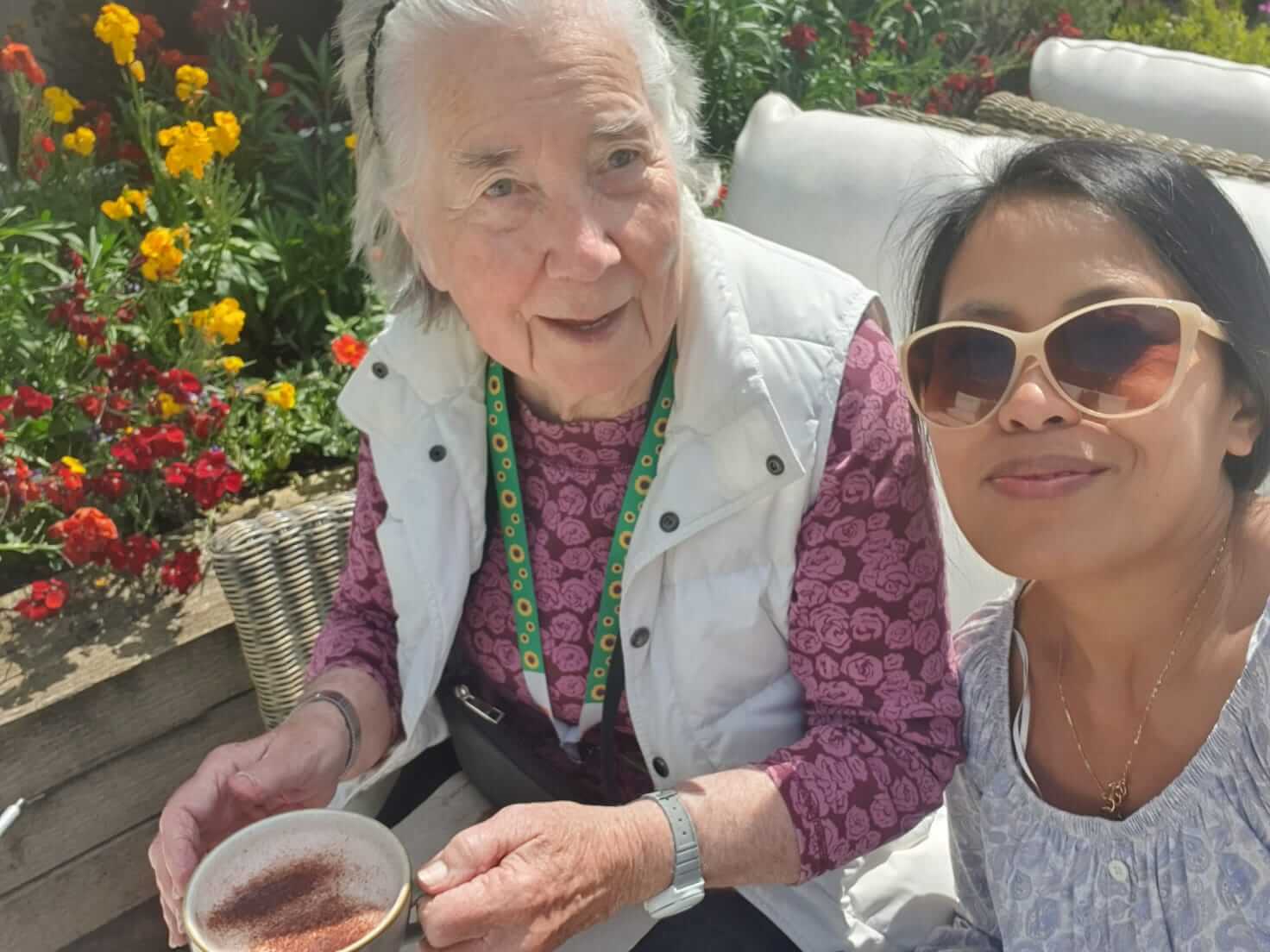 Two women sitting outside with flowers in the background, one is holding a drink and both are smiling. - Home Instead