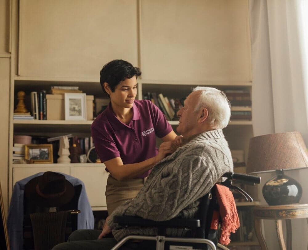 An elderly man in a wheelchair and a caregiver are talking in a living room, the man is relaxed and the caregiver smiles reassuringly. - Home Instead