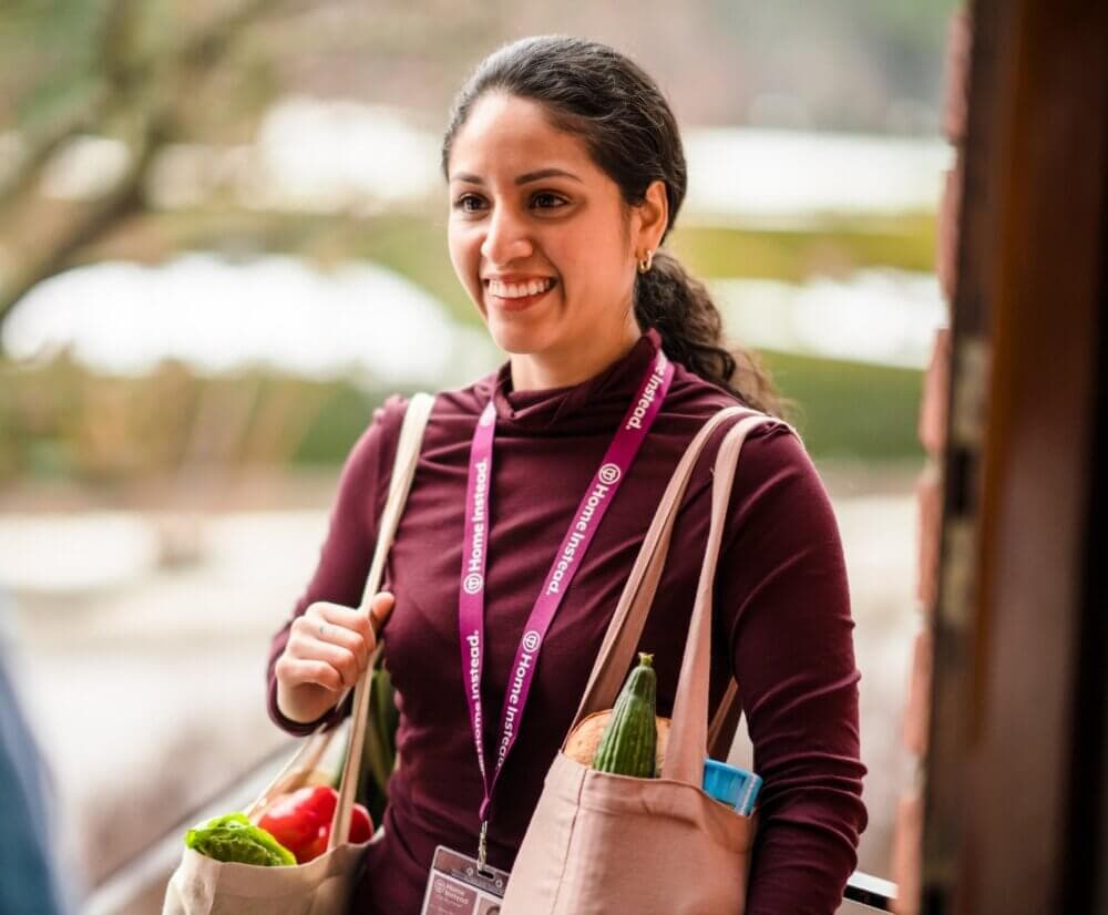 A woman smiles while carrying two bags of groceries, one with vegetables visible, standing at a doorway. - Home Instead
