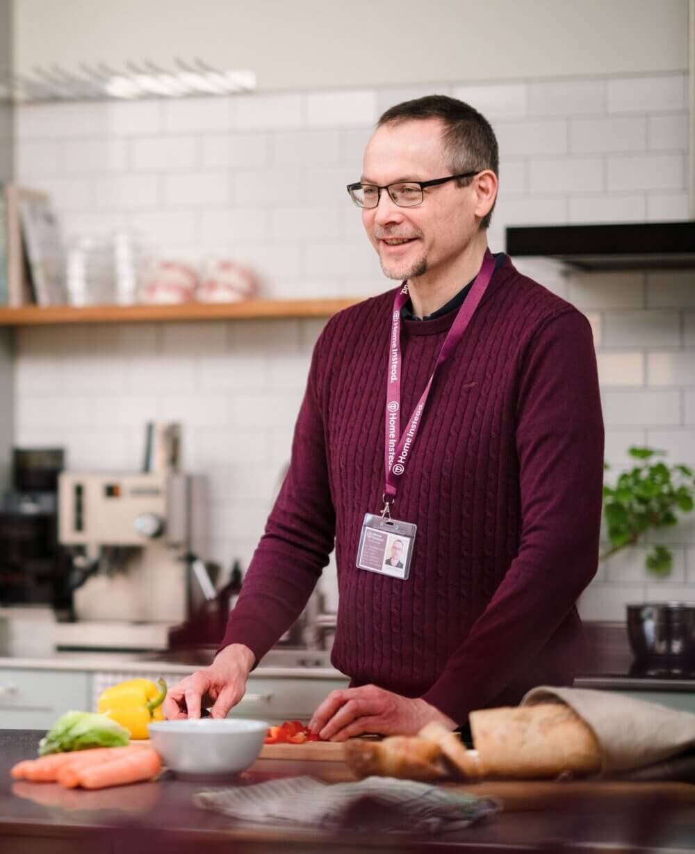 Person in a maroon sweater and glasses cooking in a modern kitchen with vegetables and bread on the counter. - Home Instead