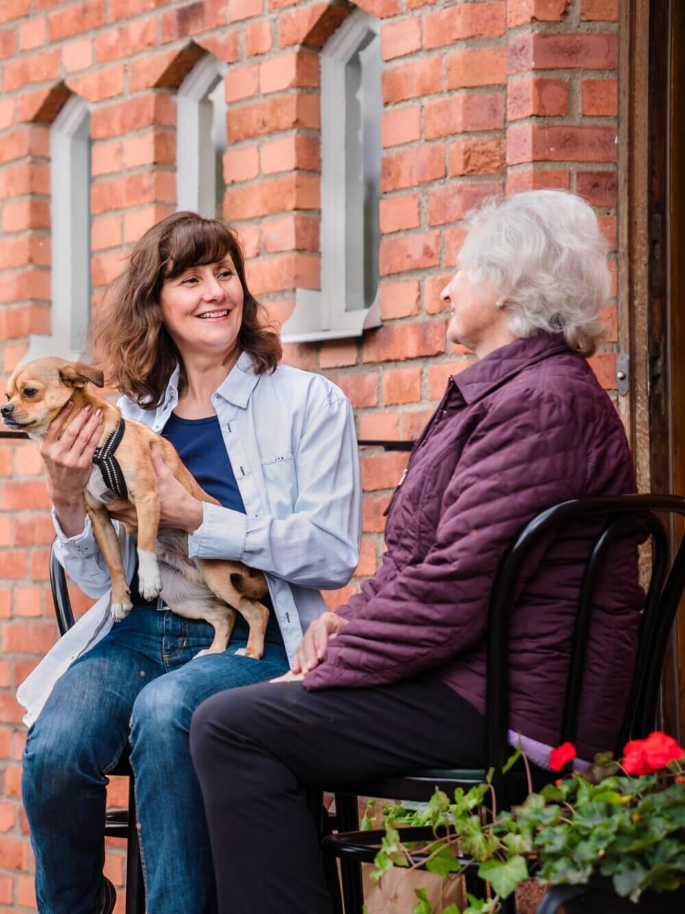 A woman holding a small dog sits next to an elderly woman on a bench outside a brick building, both smiling. - Home Instead