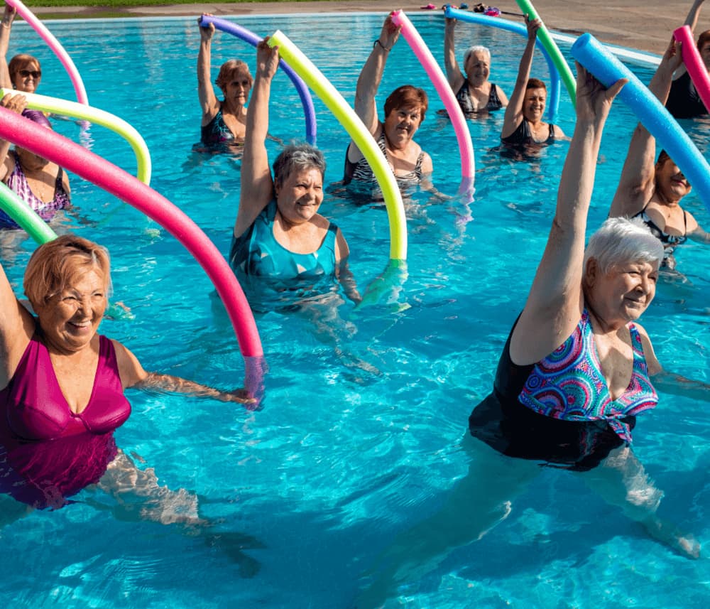 A group of elderly women in swimwear doing water aerobics with colorful pool noodles in a pool. - Home Instead