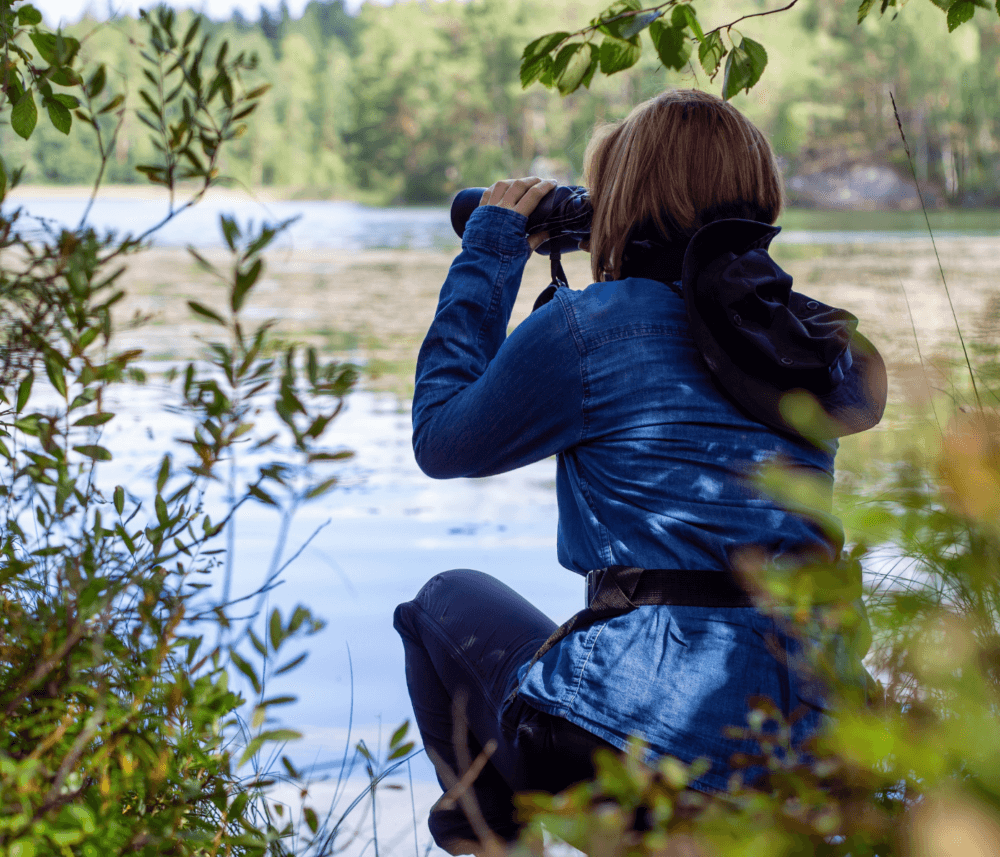 Person in a blue jacket kneeling and looking through binoculars at a lake with greenery around. - Home Instead