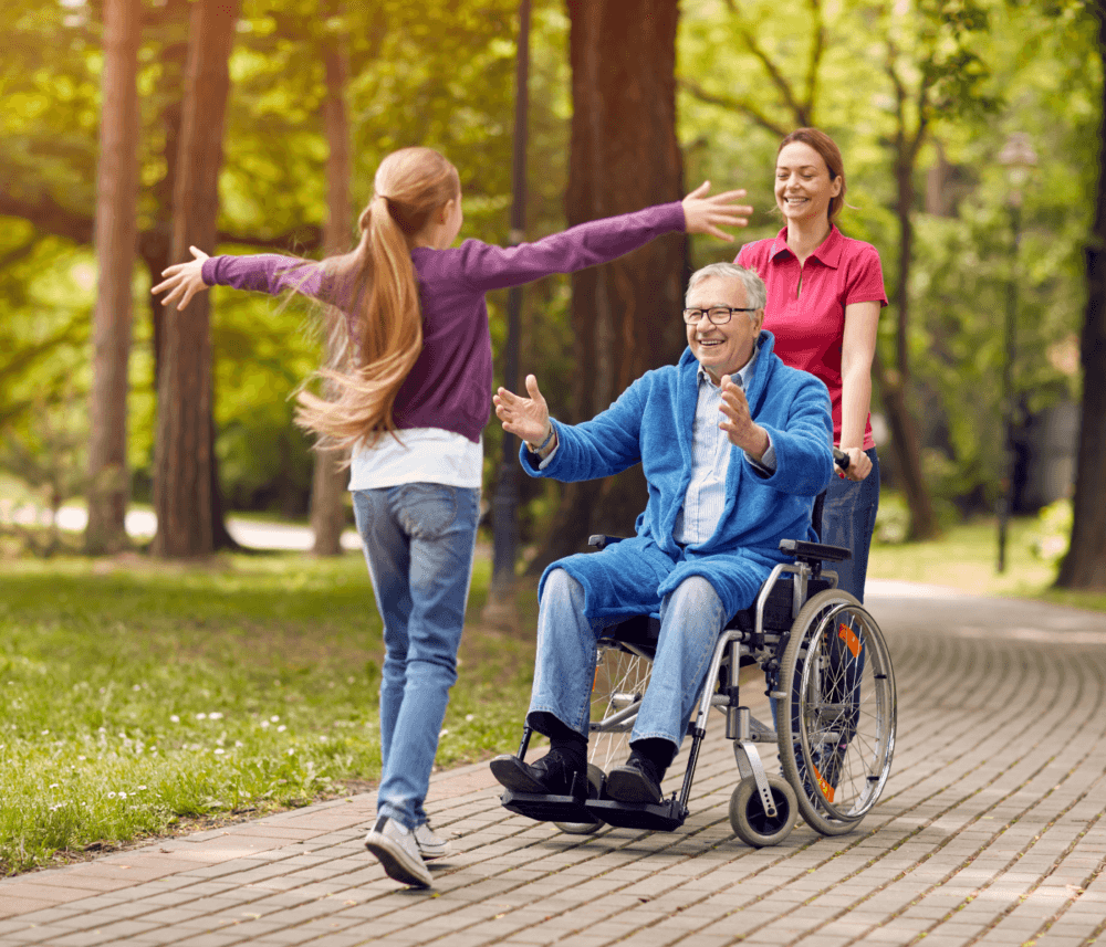 A girl runs toward a smiling elderly man in a wheelchair, pushed by a woman, in a park. - Home Instead