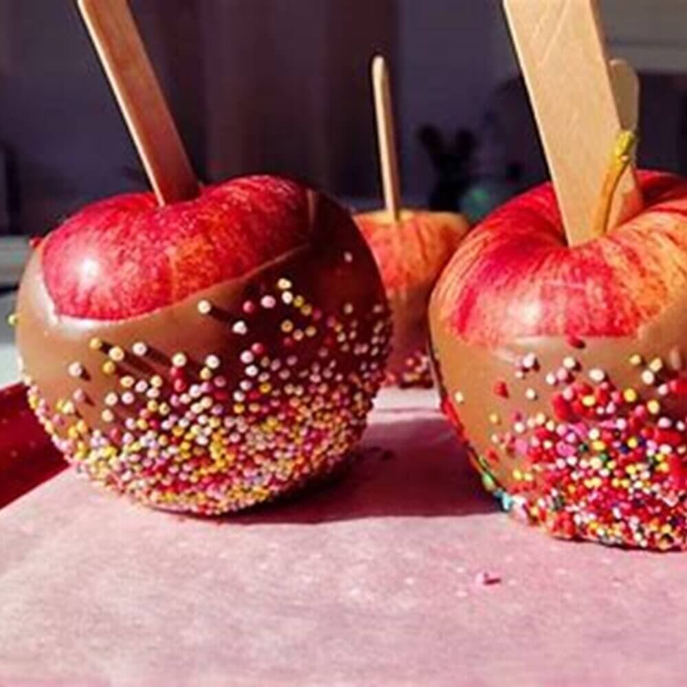 Chocolate-covered apples with sprinkles on a tray, each with a wooden stick inserted for easy holding. - Home Instead