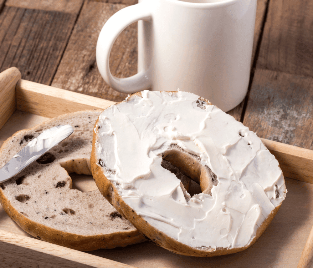 A raisin bagel with cream cheese on a wooden tray next to a white mug and a butter knife. - Home Instead