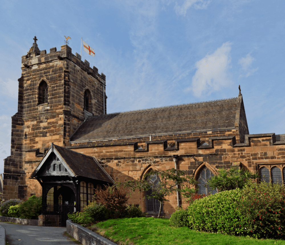 Historic stone church with a large tower, flying flags and surrounded by greenery under a partly cloudy sky. - Home Instead