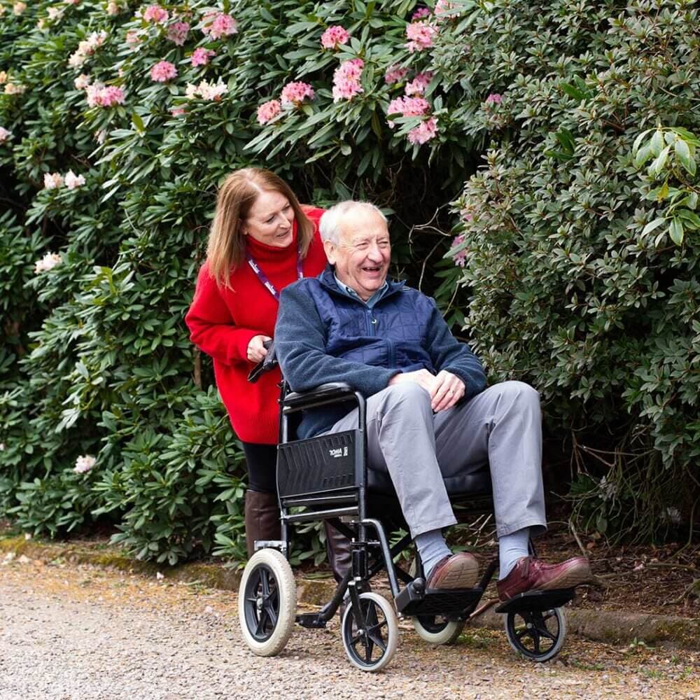 Woman in red coat pushing an elderly man in a wheelchair, both smiling, surrounded by bushes with pink flowers. - Home Instead