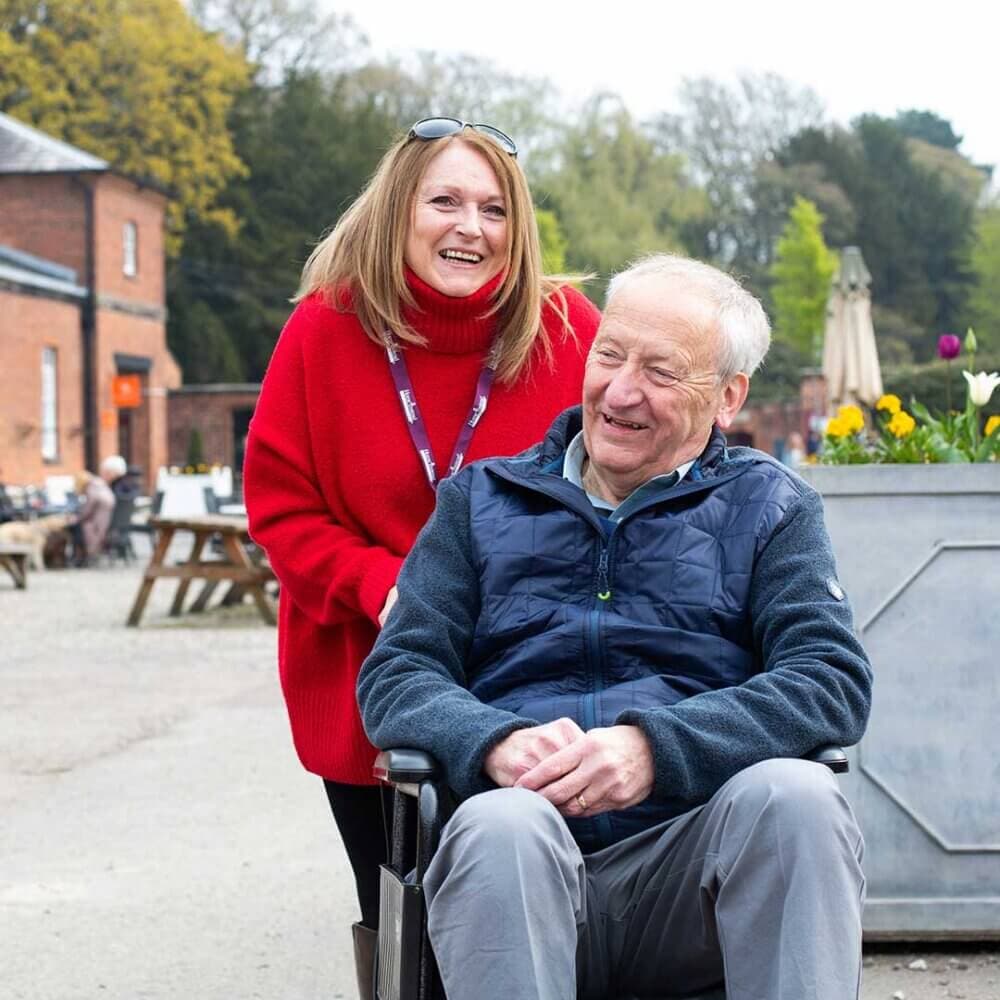 An older man in a wheelchair smiles as a woman in a red sweater stands behind him outdoors. - Home Instead