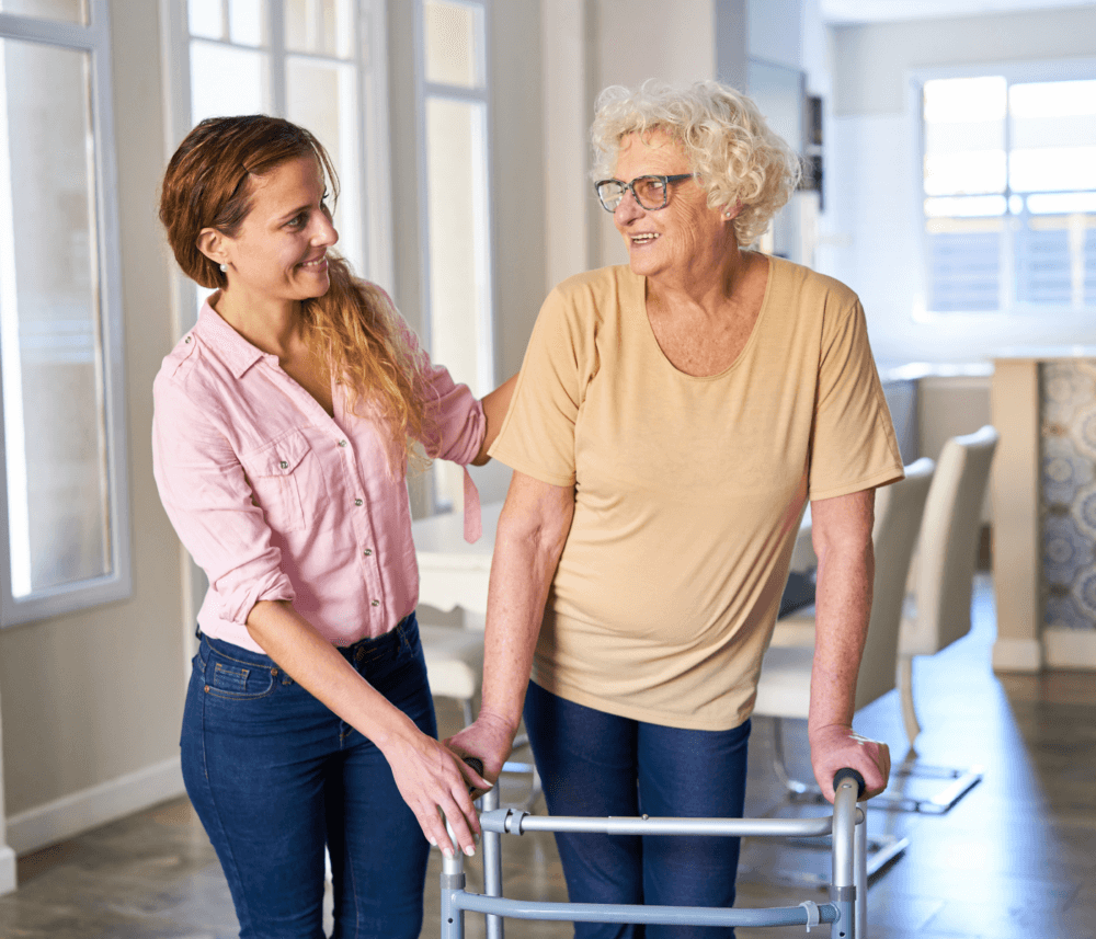 A young woman assists an older woman using a walker in a brightly lit room. Both are smiling at each other. - Home Instead
