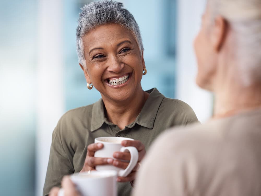 Two people enjoying a conversation over coffee indoors. One person is smiling while holding a white mug. - Home Instead