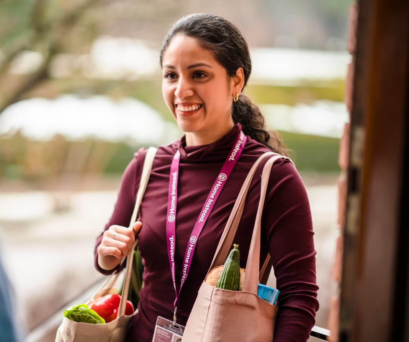 A smiling woman carrying grocery bags stands by a doorway, wearing a purple lanyard and a maroon long-sleeve shirt. - Home Instead