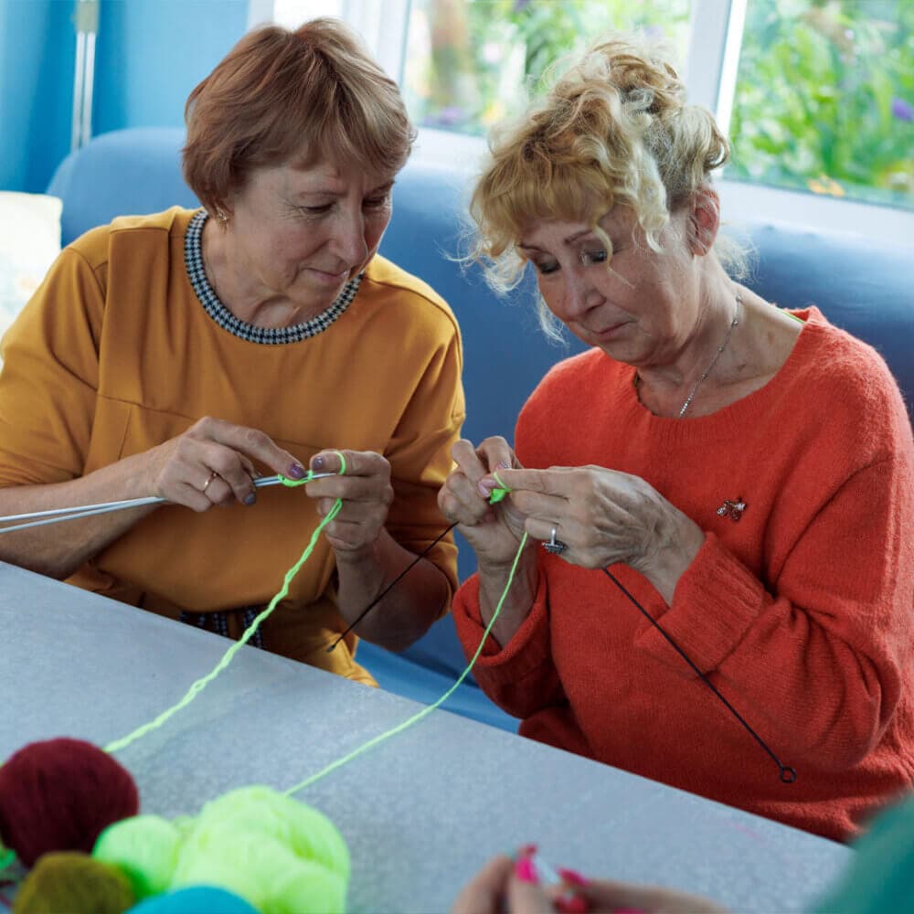 Two women sitting and knitting together, focusing on their yarn and needles, with balls of yarn on the table. - Home Instead