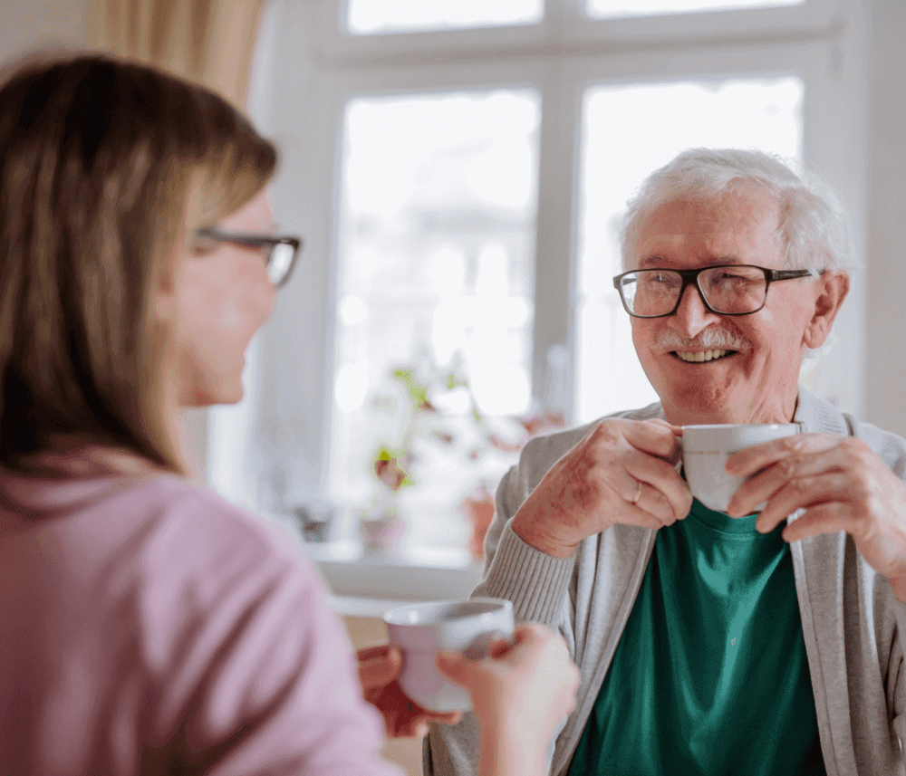 Older man and younger woman smiling and chatting while holding coffee cups in a bright room. - Home Instead
