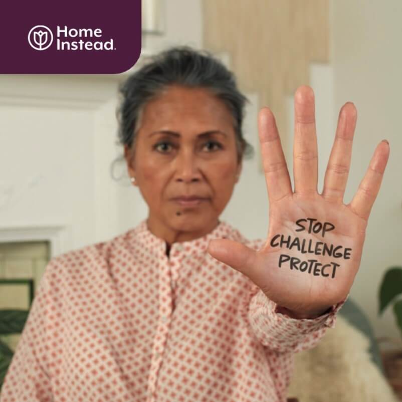An older woman holds up her hand with "STOP CHALLENGE PROTECT" written on her palm; "Home Instead" logo in corner. - Home Instead