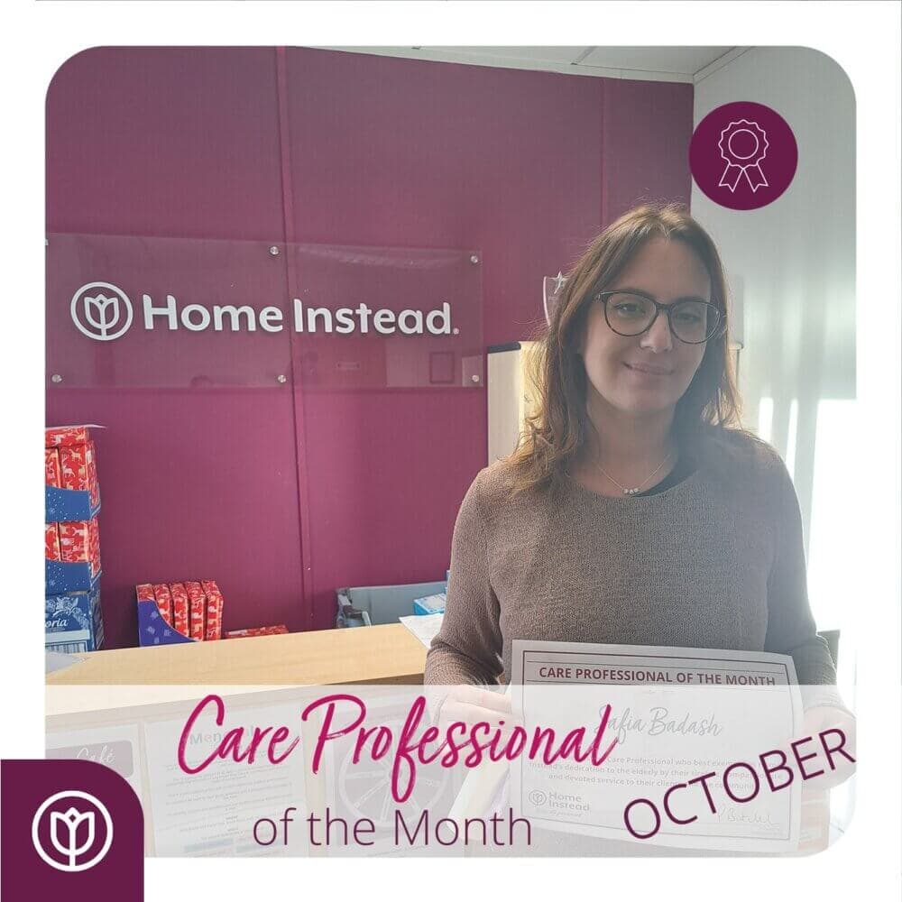 A person stands smiling in an office, holding a certificate for "Care Professional of the Month" for October. - Home Instead