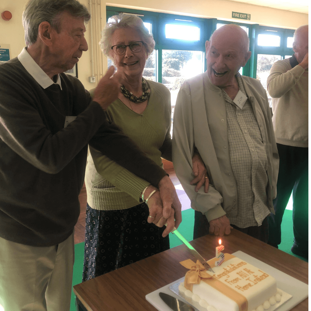 Three elderly people cutting a celebratory cake together, while smiling and conversing. - Home Instead
