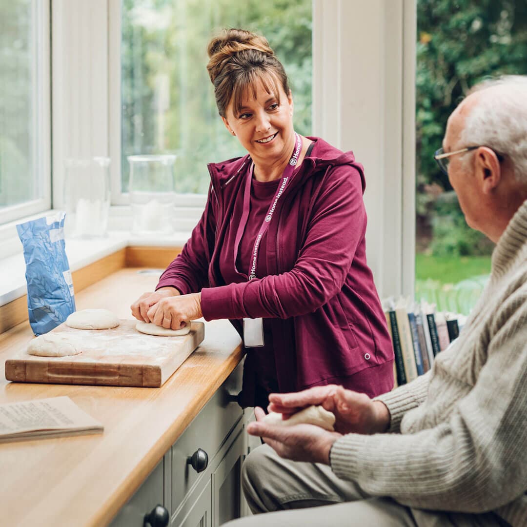 A woman kneads dough while an elderly man sits nearby holding dough, both smiling in a cozy, well-lit kitchen. - Home Instead