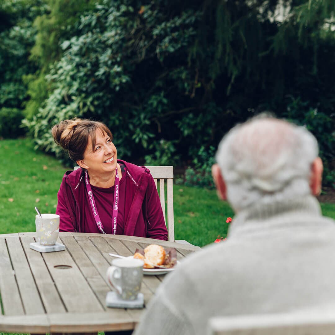 Woman in red sweater smiling at elderly man at an outdoor table with coffee. Green trees in the background. - Home Instead