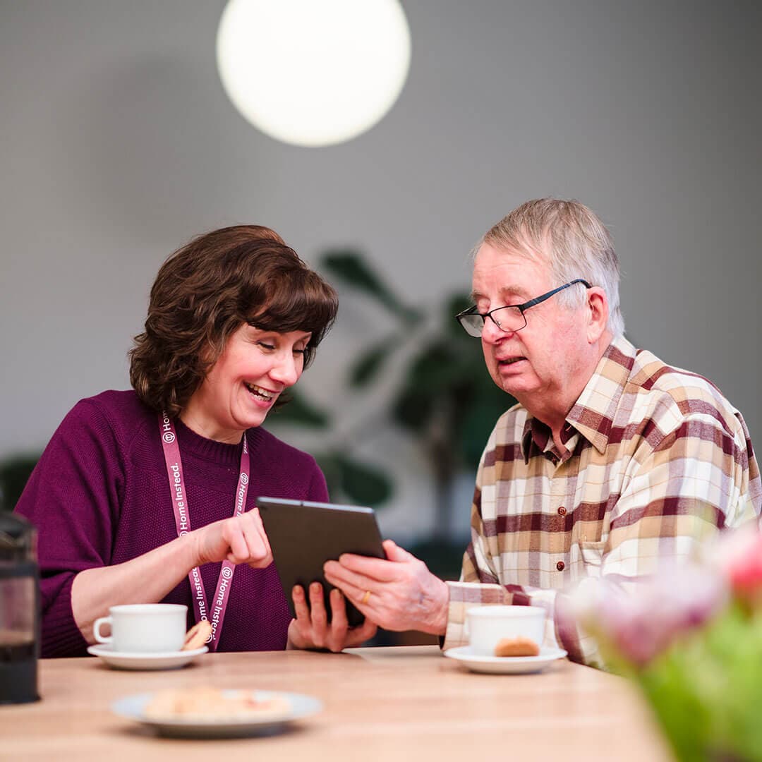 A woman helps an elderly man use a tablet at a table with coffee cups, a French press, and a blurred background. - Home Instead