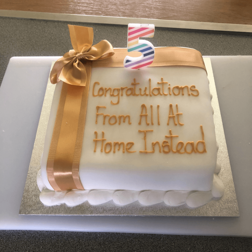 A white cake with a gold ribbon and a striped "5" topper, reading "Congratulations From All At Home Instead. - Home Instead