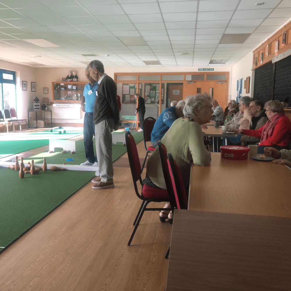 People engage in indoor lawn bowling; several sit at tables chatting, while others prepare to bowl on green mats. - Home Instead