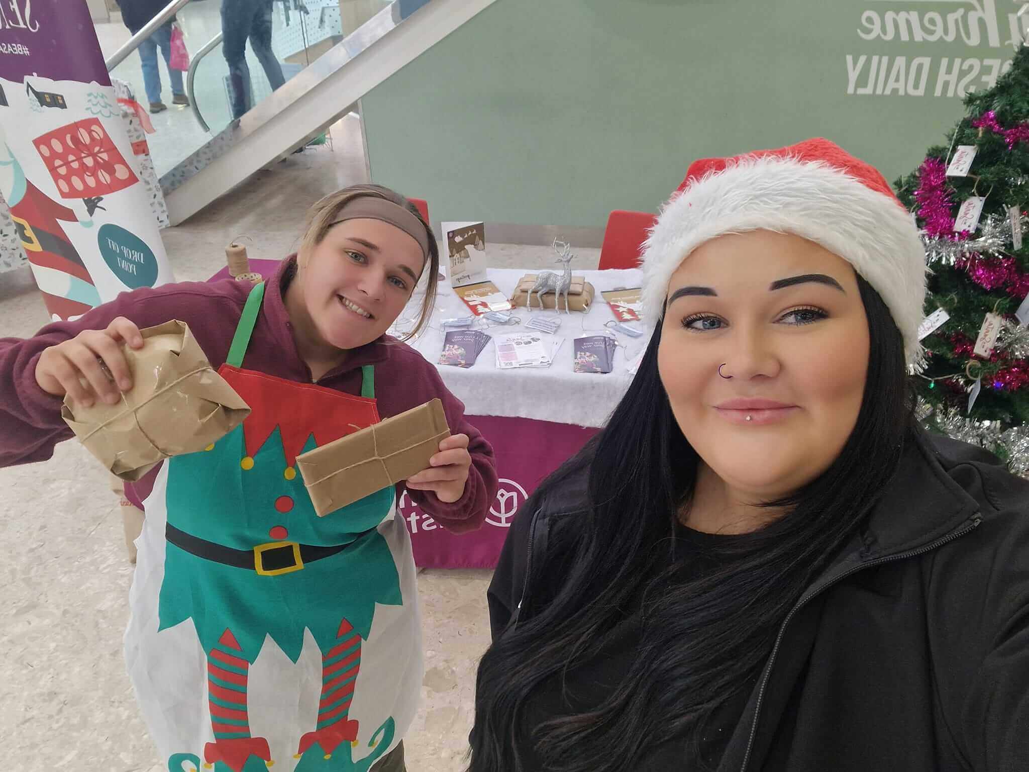 Two people smiling, one in an elf apron holding gifts, the other in a Santa hat, in a festive setting with a Christmas tree. - Home Instead