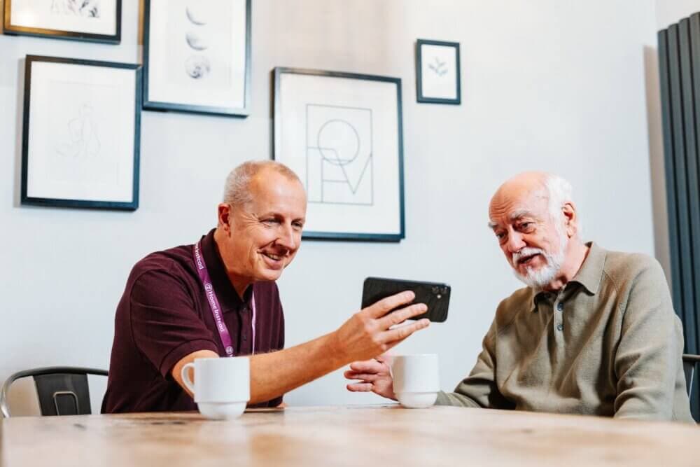 Two men sitting at a table with coffee cups, smiling and looking at a smartphone. Art frames are on the wall behind them. - Home Instead Poole