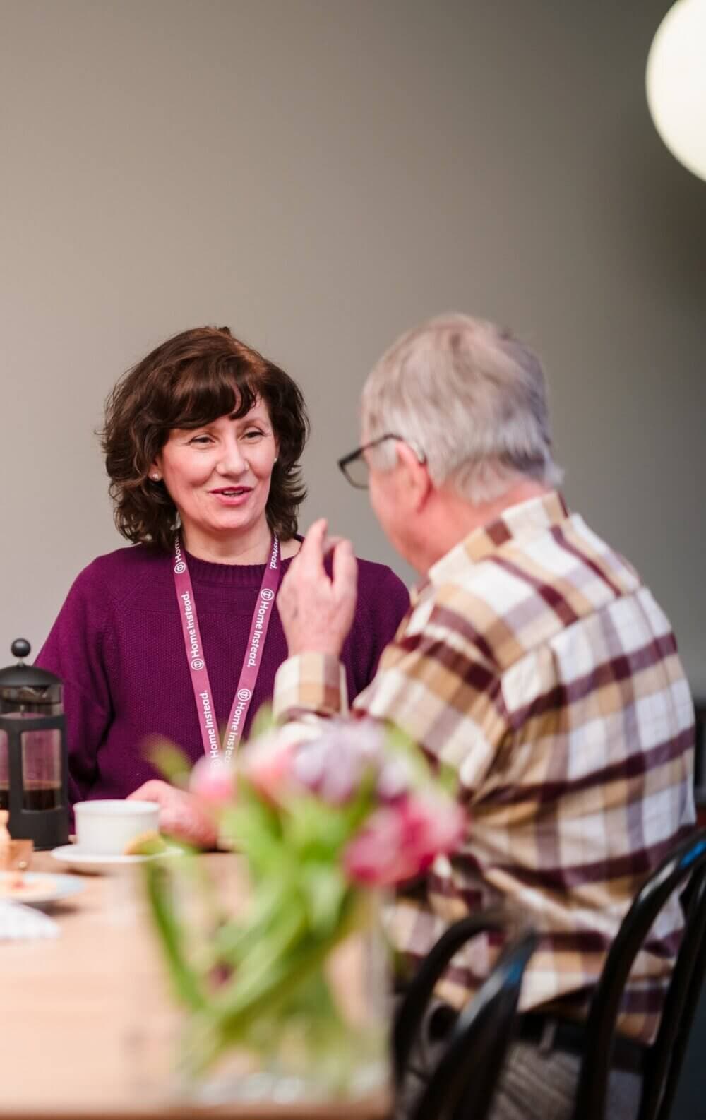 Two people engage in conversation at a table with coffee and flowers, one wearing a name badge and the other in a plaid shirt. - Home Instead