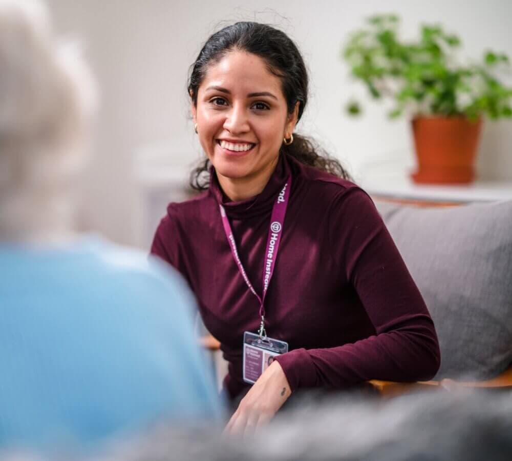 A smiling woman with dark hair wearing a maroon top and an ID badge sits with an elderly person in a light-filled room. - Home Instead