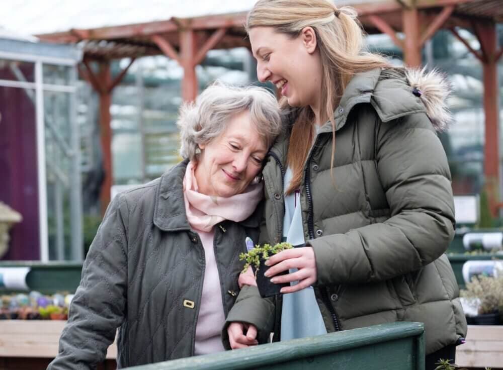 A smiling young woman and senior woman, both in jackets, share a moment while looking at plants in a greenhouse. - Home Instead