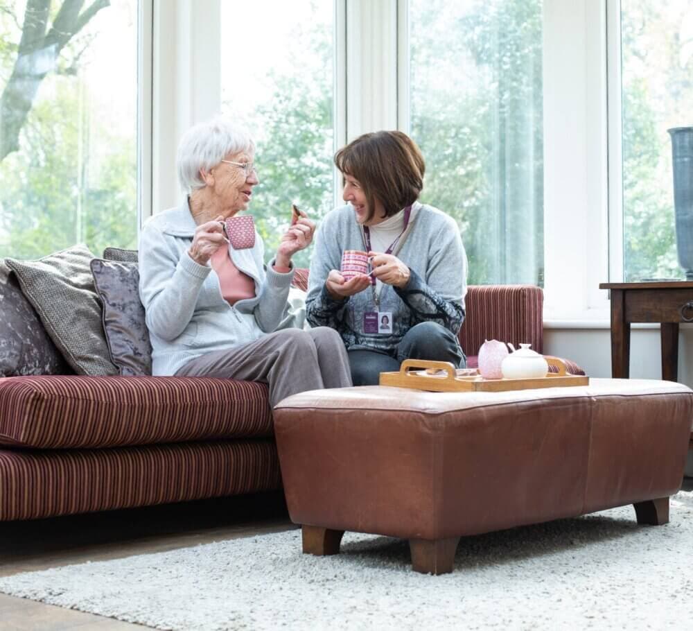 An older woman and a caregiver share a laugh while drinking tea together in a bright, cozy living room. - Home Instead