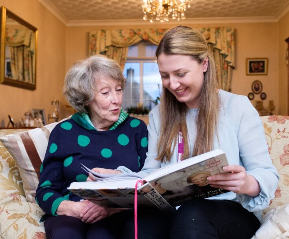 An elderly woman and a young woman sit on a couch, smiling and looking at a book together in a cozy room. - Home Instead